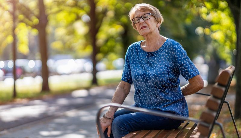 Ältere Frau, sitzt auf einer Parkbank und hält eine Hand an den unteren Rücken.