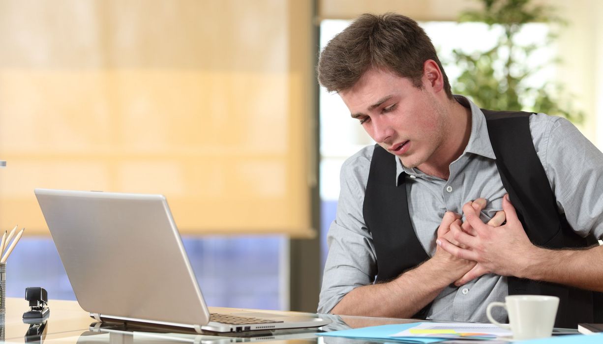 Junger Mann am Schreibtisch, offener Laptop, greift sich mit beiden Händen ans Herz.