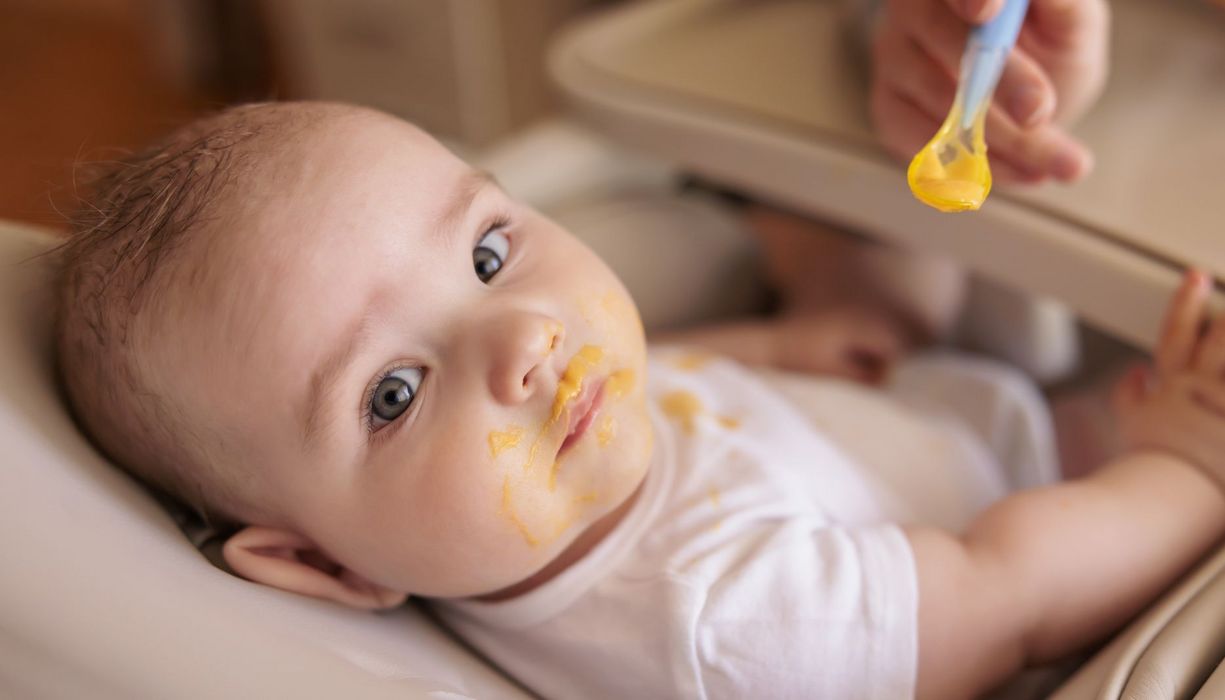 Ein etwa sechs Monate altes Baby liegt in einer Schale, der Mund ist mit orangenem Brei verschmiert. Der leere Löffel wird ihm noch hingehalten.