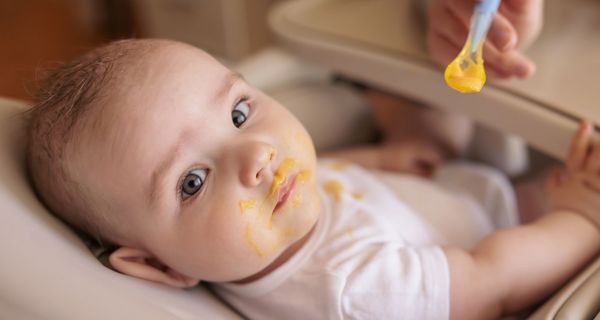 Ein etwa sechs Monate altes Baby liegt in einer Schale, der Mund ist mit orangenem Brei verschmiert. Der leere Löffel wird ihm noch hingehalten.
