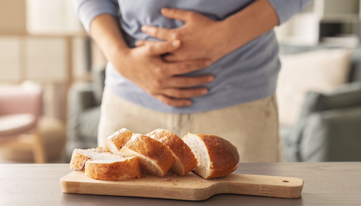 Im Vordergrund: Aufgeschnittenes Weißbrot, dahinter: Eine Person hält sich den Bauch. 
