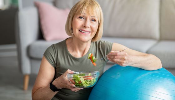 Ältere Frau, sitzt mit einem Medizinball auf dem Boden und isst Salat.