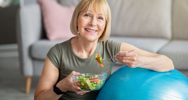 Ältere Frau, sitzt mit einem Medizinball auf dem Boden und isst Salat.
