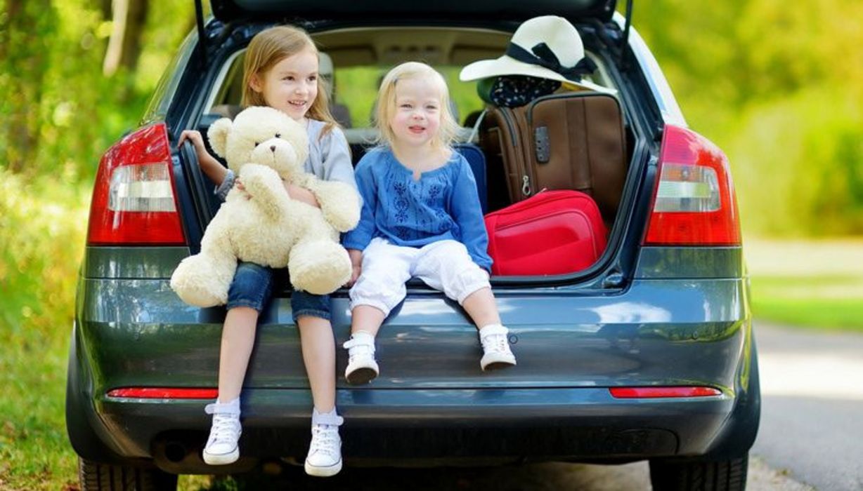 Zwei kleine Mädchen sitzen zusammen mit vielen Koffern im offenen Kofferraum eines Autos.