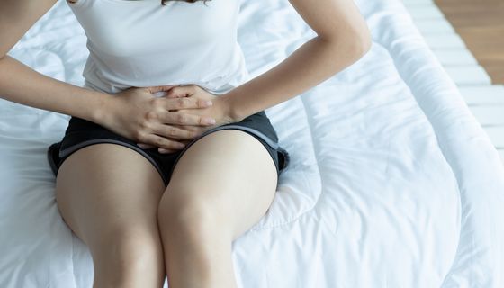 Frau, sitzt auf einem Bett und hält ihre Hände gegen den Unterleib.