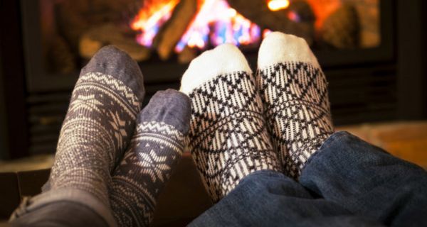 Zwei Fußpaare mit Wintersocken strecken sich einem prasselnden Kaminfeuer entgegen