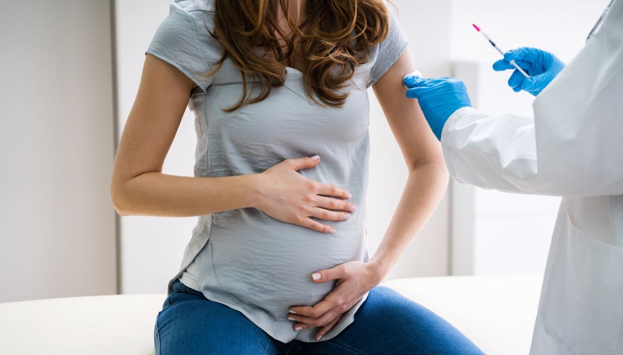 Schwangere Frau hält sich ihren Bauch und schaut Arzt mit Spritze an