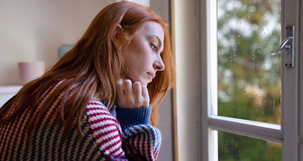 Junge Frau, sitzt vor dem Fenster und schaut traurig nach unten.