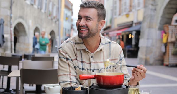 Ein Mann sitzt in einer Fußgängerzone und lächelt. Er hat eine Fonduegabel in der Hand und eine Käsefondue vor sich.