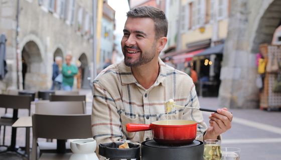 Ein Mann sitzt in einer Fußgängerzone und lächelt. Er hat eine Fonduegabel in der Hand und eine Käsefondue vor sich.