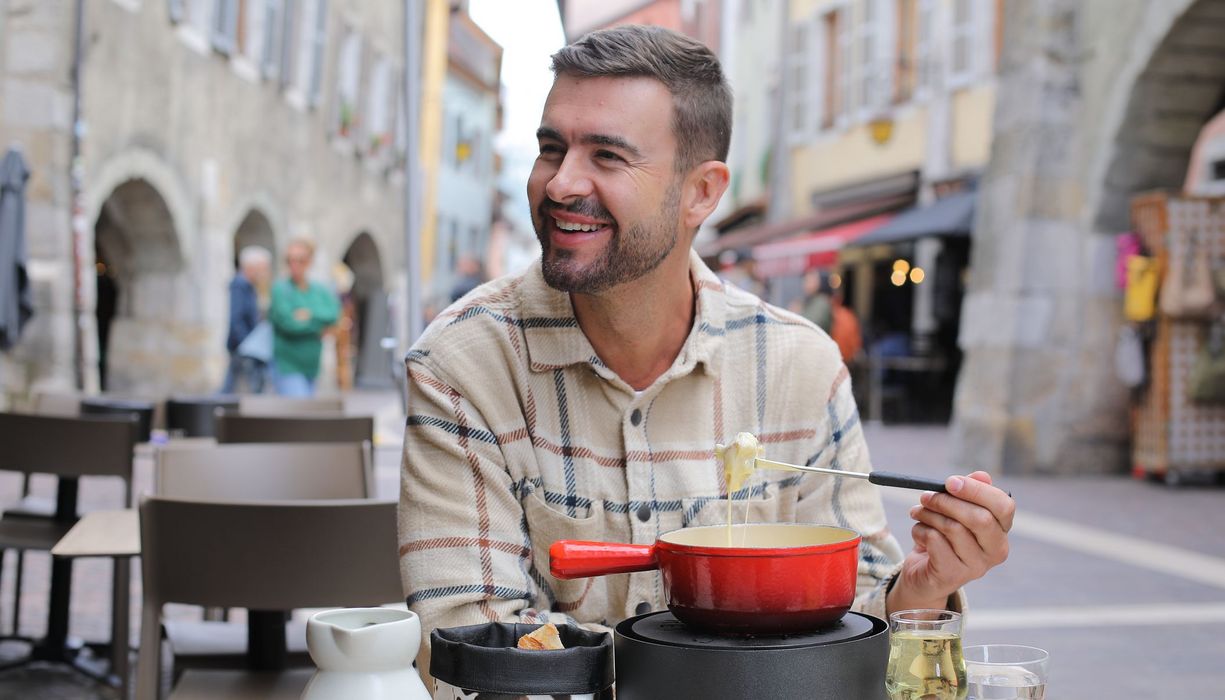 Ein Mann sitzt in einer Fußgängerzone und lächelt. Er hat eine Fonduegabel in der Hand und eine Käsefondue vor sich.