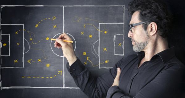 Fußballtrainer, schwarzes Hemd, an Tafel eine Strategie mit gelbem Stift auf Tafel zeichnend (steht am rechten Bildrand, Profil, etwas der Tafel zugewandt)