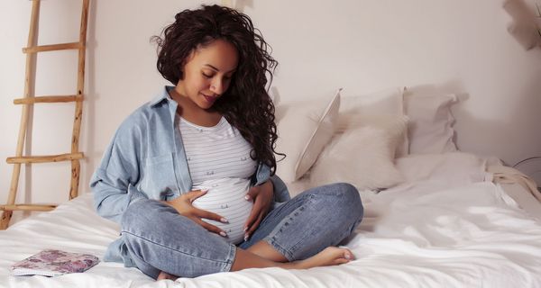 Junge schwangere Frau, sitzt im Schneidersitz auf dem Bett und hält ihren Bauch.