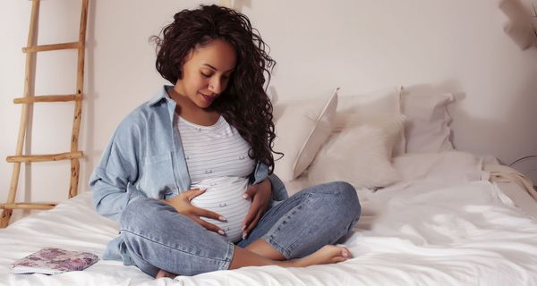 Junge schwangere Frau, sitzt im Schneidersitz auf dem Bett und hält ihren Bauch.