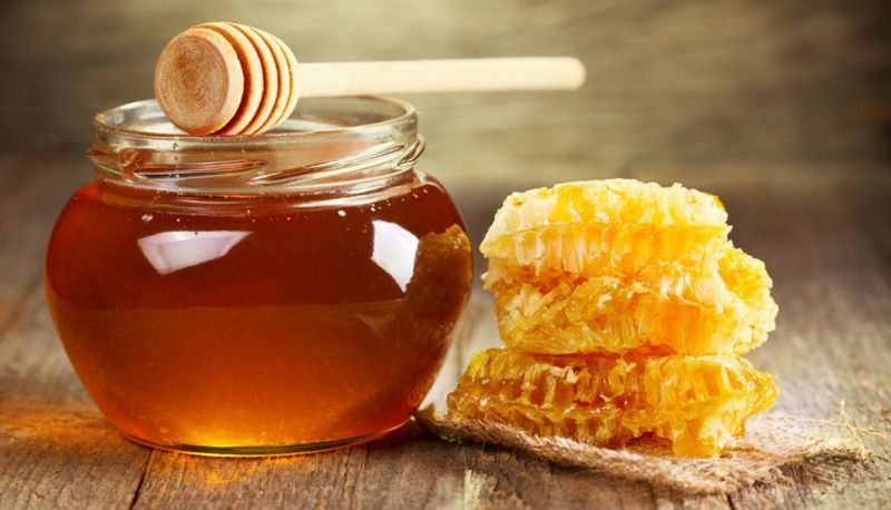 Auf Holztisch: rundliches Schraubglas mit Honig, darauf Honigspindel, daneben Honigwaben