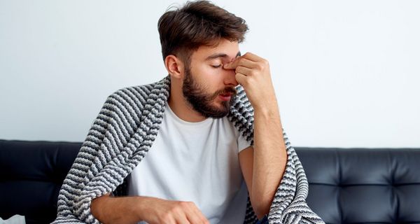 Mann mit Nasennebenhöhlenentzündung sitzt mit Decke und Taschentuchspender auf einer Couch. Er hält sich den Nasenrücken.