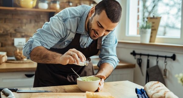 Junger Mann, steht in der Küche und benutzt einen Salzspender, um Guacamole zu würzen.