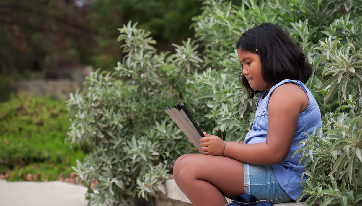 Junges Mädchen, sitzt draußen und schaut in einen Laptop.