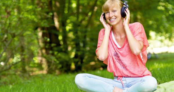 Junge Frau sitzt im Schneidersitz auf einer Wiese und hört glücklich Musik über Kopfhörer