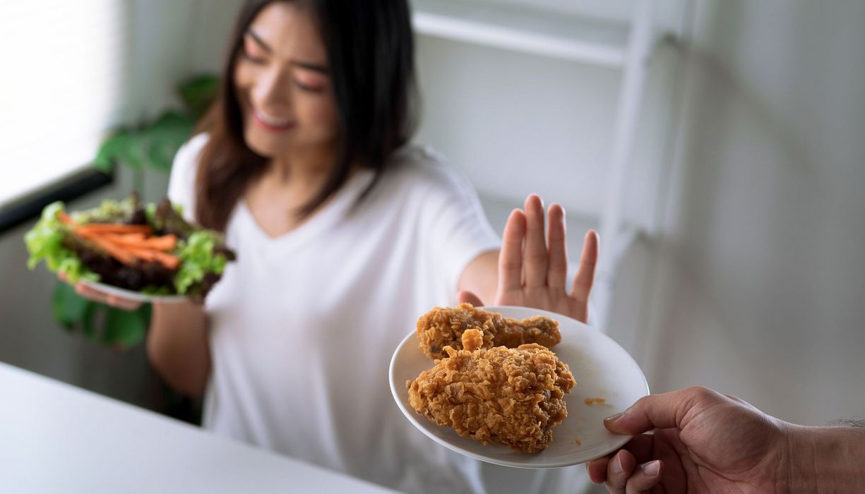 Eine junge Frau lehnt einen Teller mit frittiertem Hühnchen ab. Sie blickt lächelnd auf einen Teller mit Salat und Karottenstreifen.