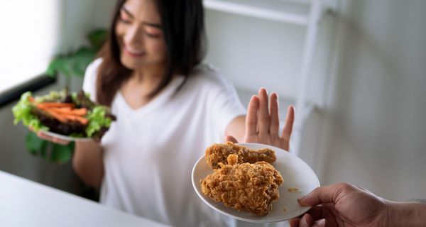Eine junge Frau lehnt einen Teller mit frittiertem Hühnchen ab. Sie blickt lächelnd auf einen Teller mit Salat und Karottenstreifen.