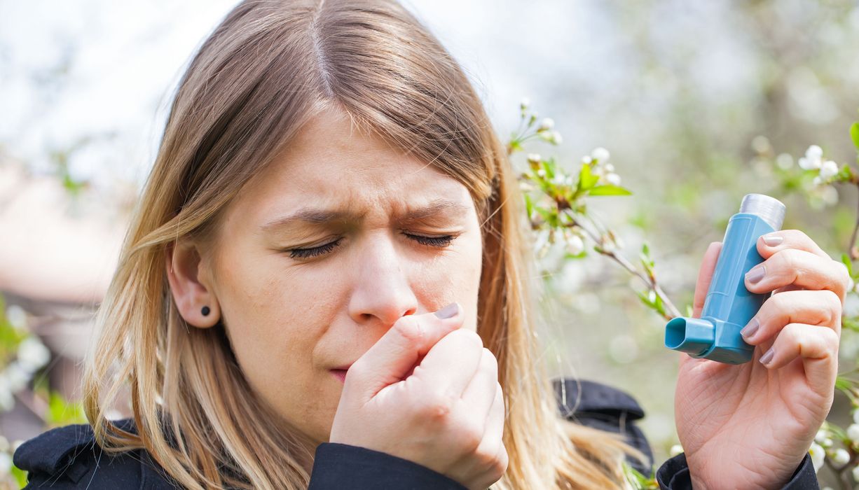 eine Frau steht vor einem blühenden Baum, hustet und hat ein Asthma-Spray in der Hand