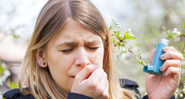 eine Frau steht vor einem blühenden Baum, hustet und hat ein Asthma-Spray in der Hand