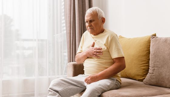 Opa sitzt auf seinem Sofa und fasst sich angespannt mit der rechten Hand an die Brust.