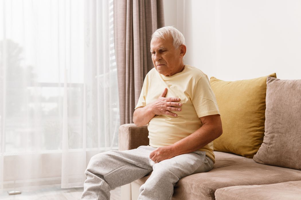 Opa sitzt auf seinem Sofa und fasst sich angespannt mit der rechten Hand an die Brust.