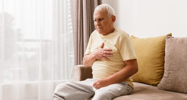 Opa sitzt auf seinem Sofa und fasst sich angespannt mit der rechten Hand an die Brust.