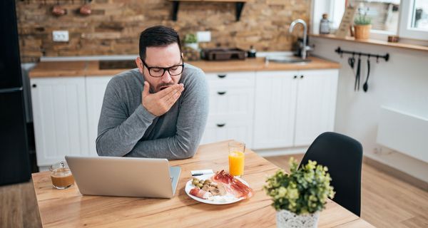 Junger Mann, sitzt mit seinem Laptop und einem Teller mit Essen in der Küche und gähnt.