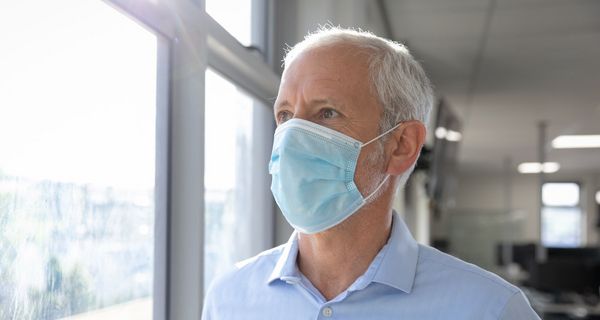 Älterer Mann mit Maske blickt aus dem Fenster.