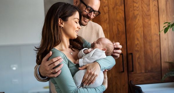 Eine Frau hält ein Baby, ein Mann hat seine Arme um sie gelegt und lächelt das Kind an.