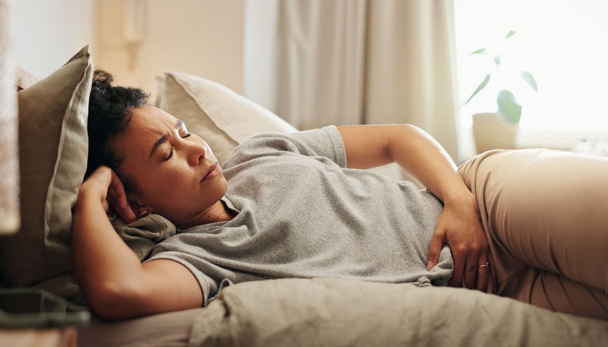 Frau hält sich den Unterbauch und liegt im Bett. Sie wirkt angespannt.