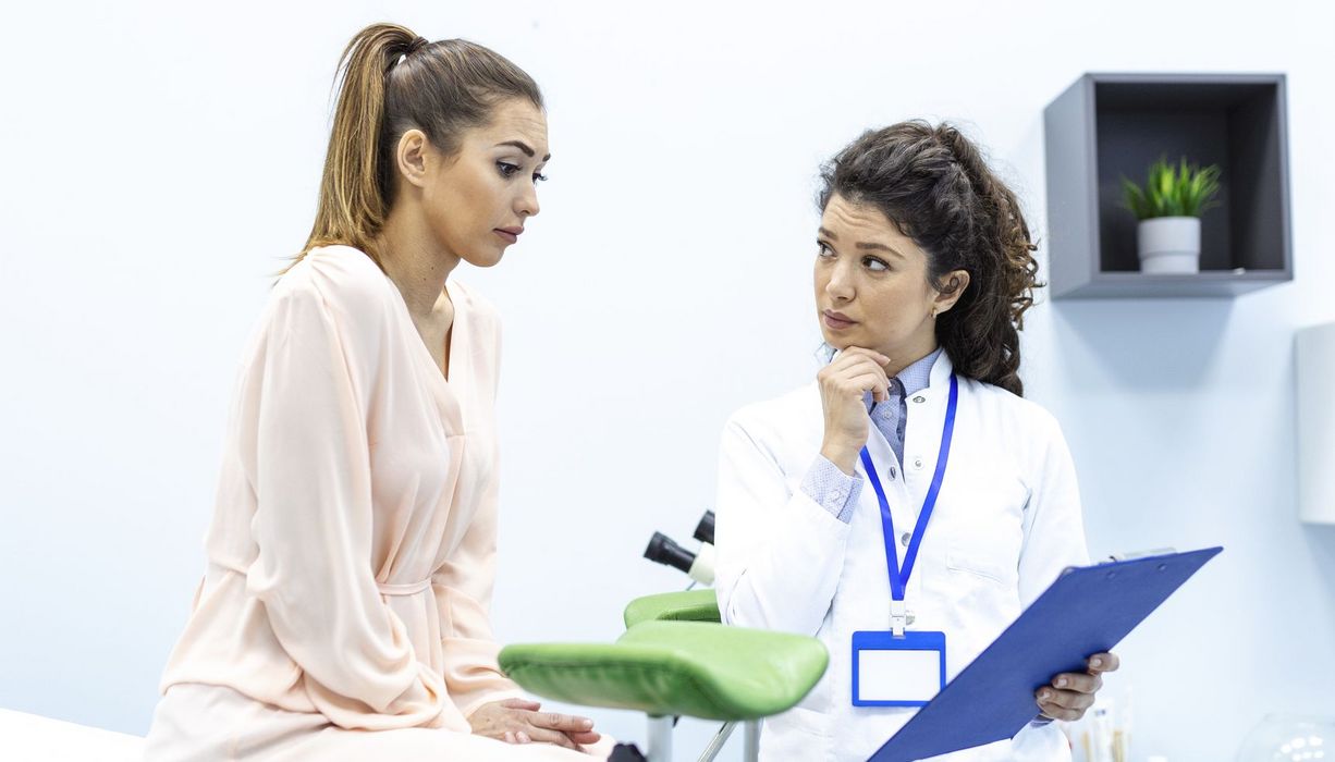 Eine Frau sitzt auf dem Untersuchungstisch bei der Gynäkologin und blickt unsicher auf ein Klemmbrett. Die Ärztin schaut fragend zu ihr.