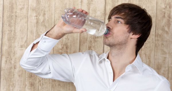 Junger dunkelhaariger Mann im hellen Hemd vor einer Holzwand trinkt Wasser aus einer Plastikflasche