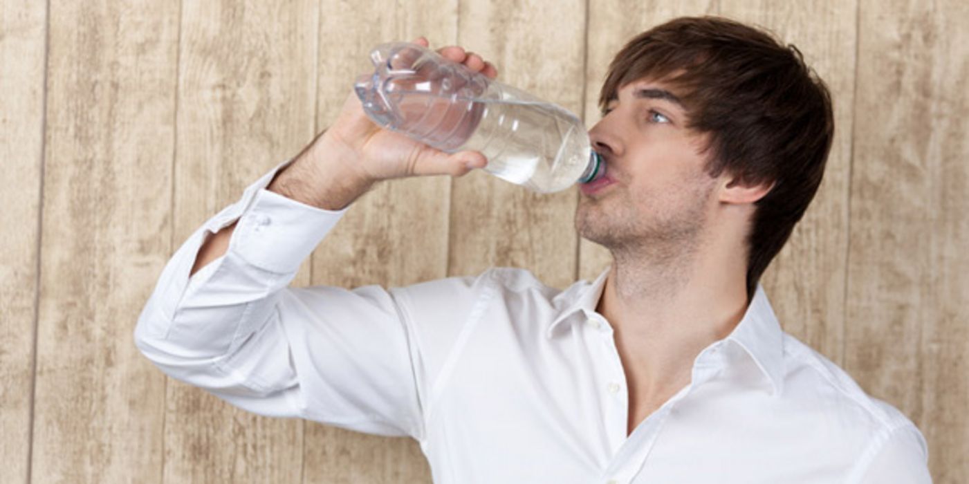 Junger dunkelhaariger Mann im hellen Hemd vor einer Holzwand trinkt Wasser aus einer Plastikflasche