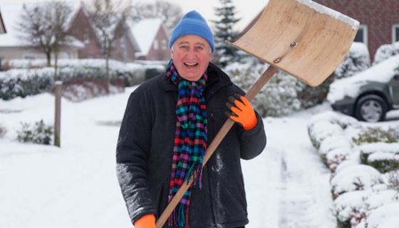 Winterszene: In die Kamera lachender Mann im Schnee, um die 60, hellblaue Pudelmütze, rosiges Gesicht, Winterjacke, bunter Schal, in den Händen Schneeschippe, die er nach oben hält. Im Hintergrund Einfamilienhäuser
