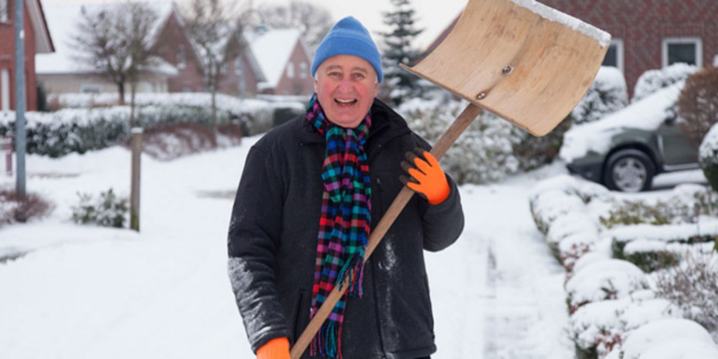 Winterszene: In die Kamera lachender Mann im Schnee, um die 60, hellblaue Pudelmütze, rosiges Gesicht, Winterjacke, bunter Schal, in den Händen Schneeschippe, die er nach oben hält. Im Hintergrund Einfamilienhäuser
