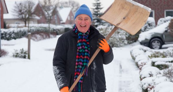 Winterszene: In die Kamera lachender Mann im Schnee, um die 60, hellblaue Pudelmütze, rosiges Gesicht, Winterjacke, bunter Schal, in den Händen Schneeschippe, die er nach oben hält. Im Hintergrund Einfamilienhäuser