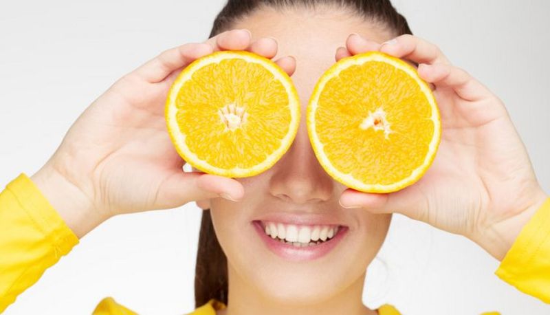 Gesicht frontal: junge Frau hält sich je eine halbe Orange oder Grapefruit (?) vor jedes Auge; Schnittfläche nach außen. Knallgelbe Bluse