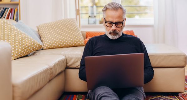 Älterer Mann sitzt auf dem Boden mit Laptop auf seinem Schoß.