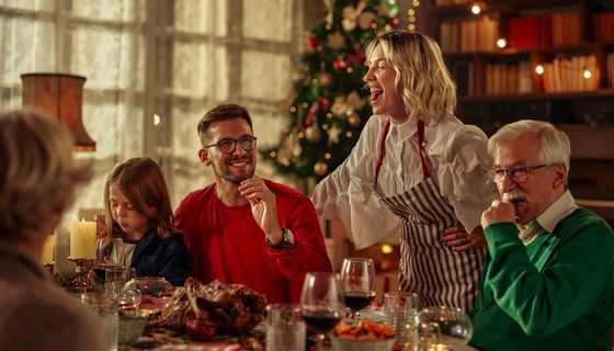 Weihnachtsessen: Eine Familie sitzt rund um den gedeckten Tisch, eine Frau steht lachend. Auf dem Tisch ist ein gebratener Vogel, Gemüse, gut gefüllte Weingläser; Im Hintergrund ein Weihnachtsbaum.
