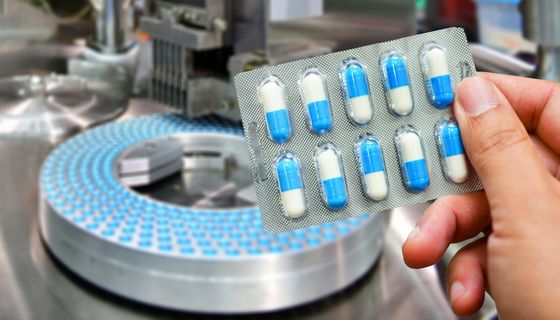 Eine Hand hält eine Blisterpackung mit blauen Kapseln, im Hintergrund werden weitere Kapseln produziert.