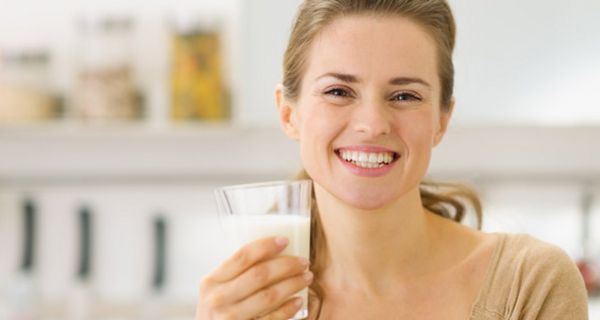 Junge, in die Kamera lachende Frau, die langen Haare zurückgebunden, mit einem Glas Milch in der linken Hand