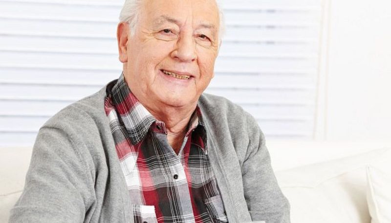 Senior, weißer Haarkranz, rot-weiß-schwarz kariertes Hemd, darüber graue Strickjacke, Tablette auf dem linken Handteller, Wasserglas in der rechten Hand, lächelt in die Kamera