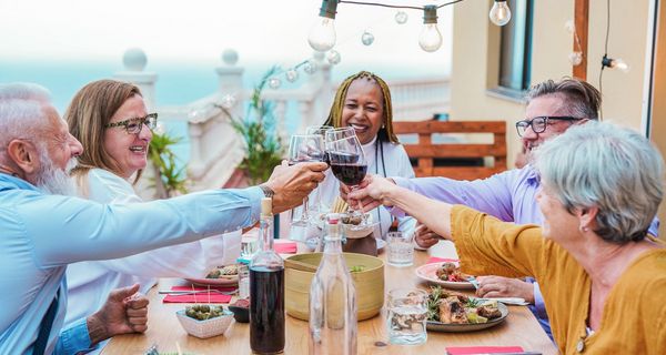 Freunde im mittleren Alter sitzen an einem gedeckten Tisch und prosten sich mit Rotwein zu. zu.