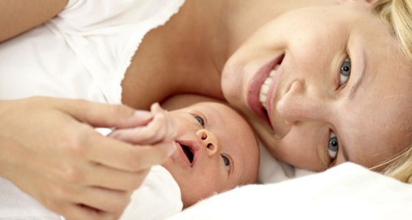 Junge Mutter mit Neugeborenem, beide auf Bett liegend, schaut lächelnd in die Kamera
