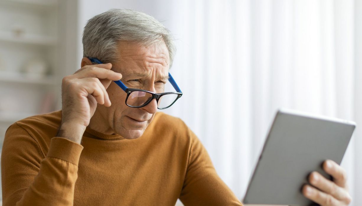 Älterer Mann versucht, auf einem Tablet etwas zu lesen. Er passt die Position der Brille an.