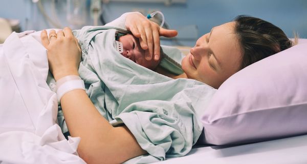 Frau liegt nach der Geburt mit ihrem Neugeborenen im Krankenhaus im BNett. Sie streichelt lächelnd die Stirn des Babys.